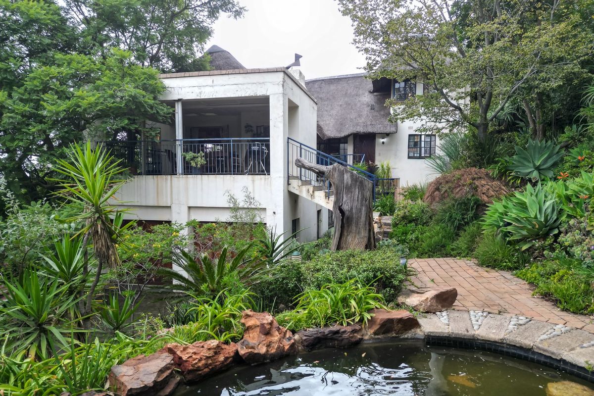 Rear view of a multi-level home featuring a thatched roof, elevated balcony, external staircase, and a landscaped garden with a pond.
