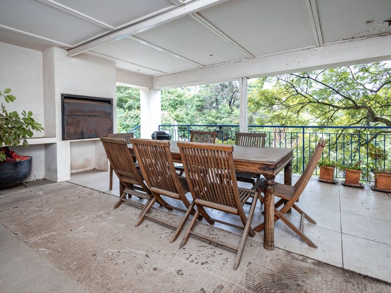 A covered outdoor patio featuring a built-in braai, tiled flooring, and metal railings, overlooking a lush garden setting.