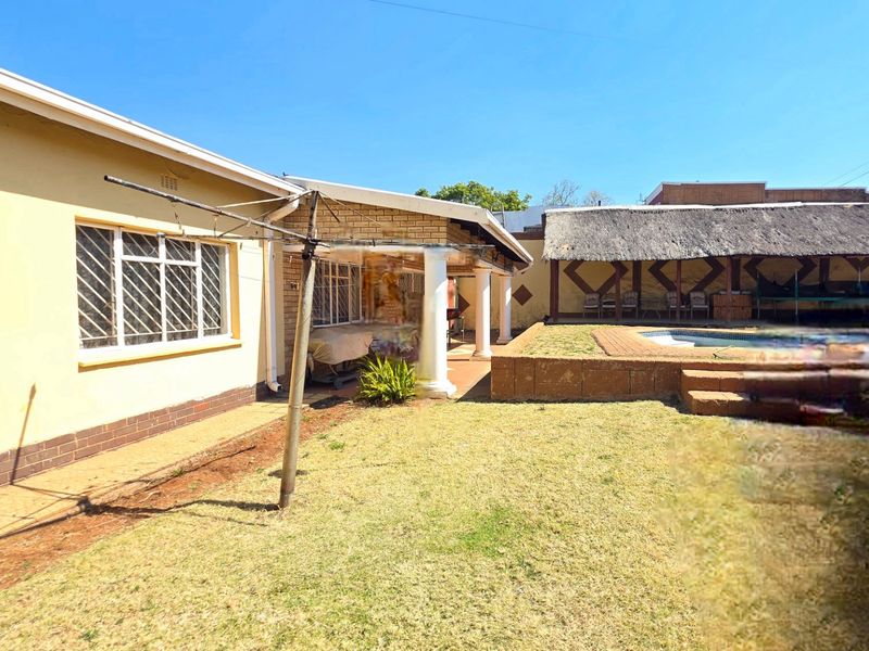 Rear garden area featuring a swimming pool, a covered patio with columns, and a traditional thatched lapa structure.