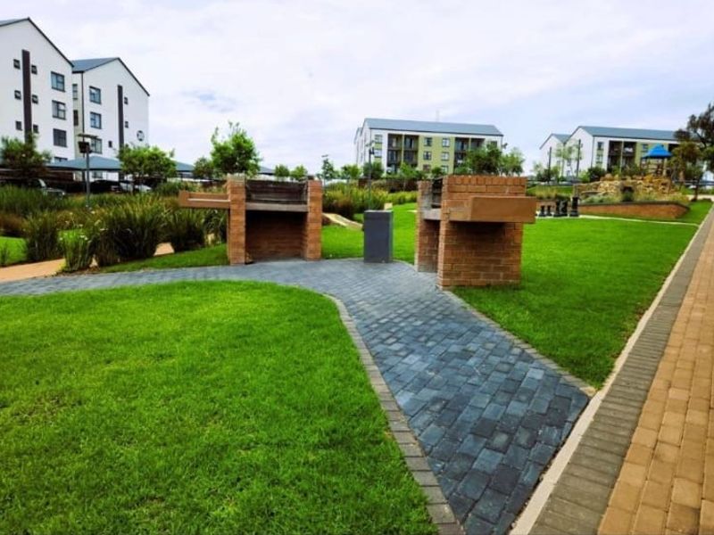 A well-maintained communal garden area featuring brick braai stands, paved pathways, and manicured lawns within an apartment complex.