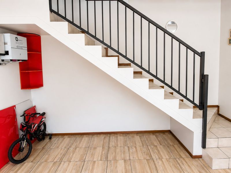 A bright hallway featuring a staircase with a modern metal balustrade, tiled flooring, and a wall-mounted inverter system.