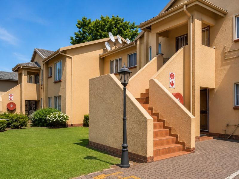Exterior view of a well-maintained apartment complex featuring a paved entrance, external staircase, landscaped lawn, and functional building infrastructure.