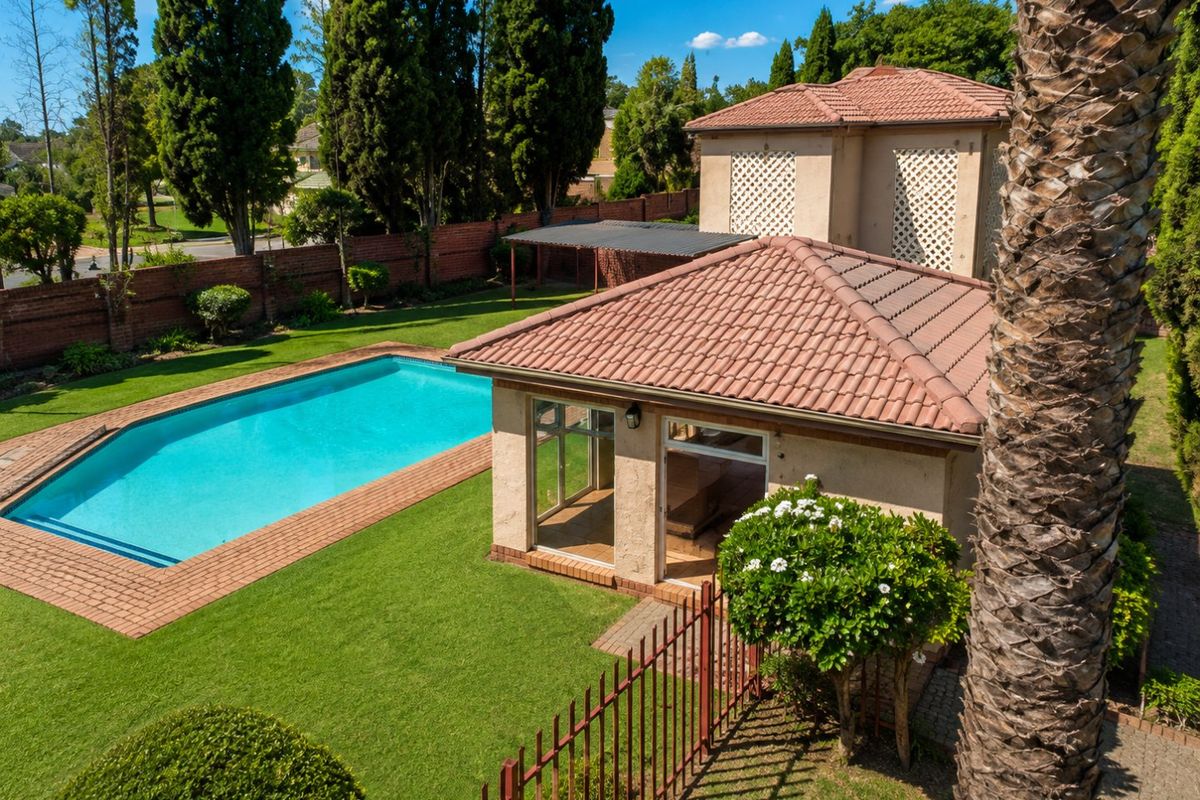 Aerial view of a well-maintained backyard featuring a swimming pool, paved surround, lush lawn, and a detached structure with a tiled roof.