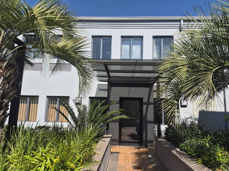 Front facade of a commercial building featuring a modern glass entrance canopy, aluminium windows, and a paved walkway surrounded by landscaped garden beds.