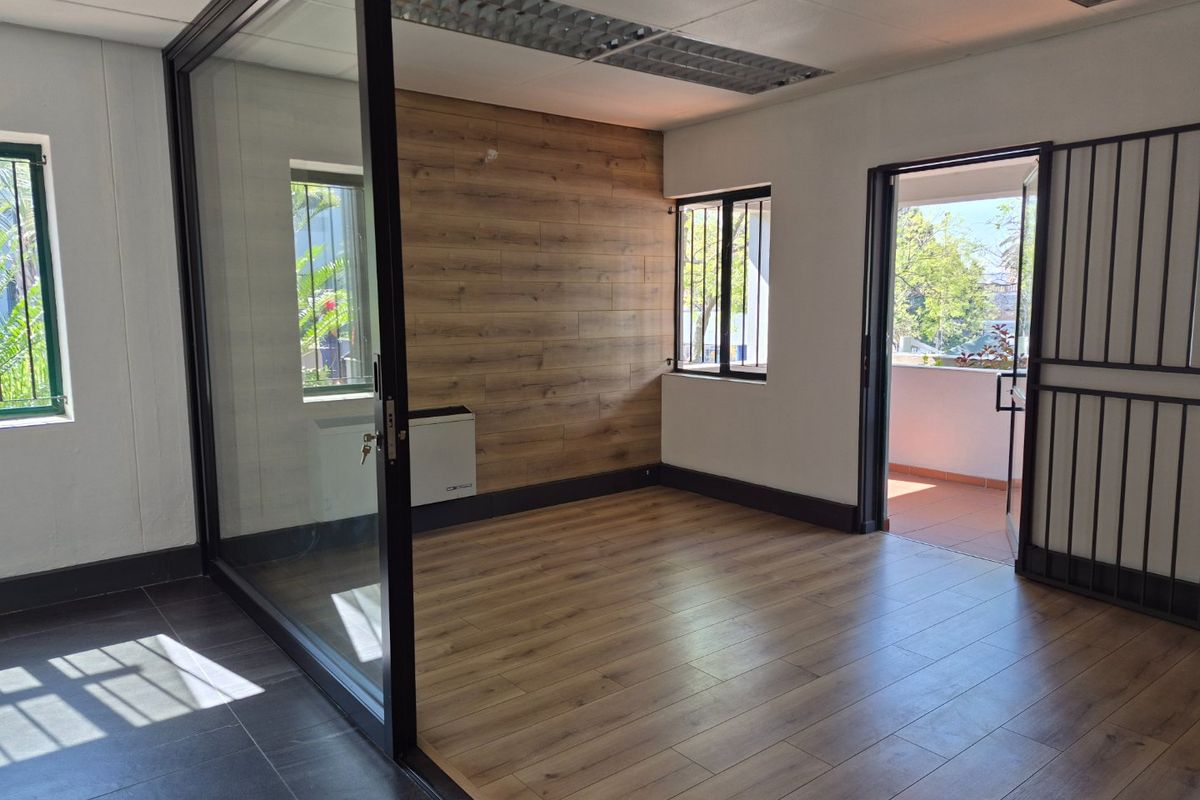 A bright, empty office space featuring wood-look flooring, a glass partition, wood-paneled accent wall, and direct access to an outdoor balcony.