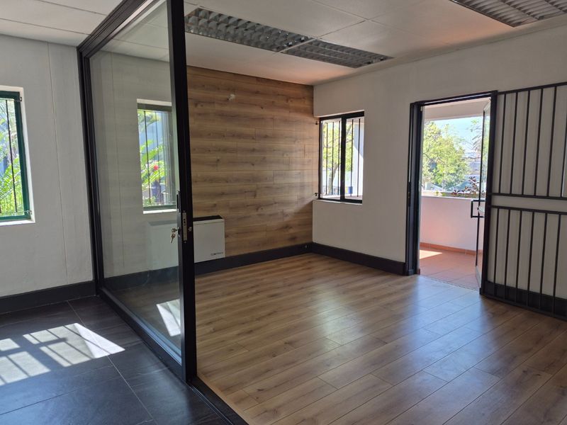A bright, empty office space featuring wood-look flooring, a glass partition, wood-paneled accent wall, and direct access to an outdoor balcony.