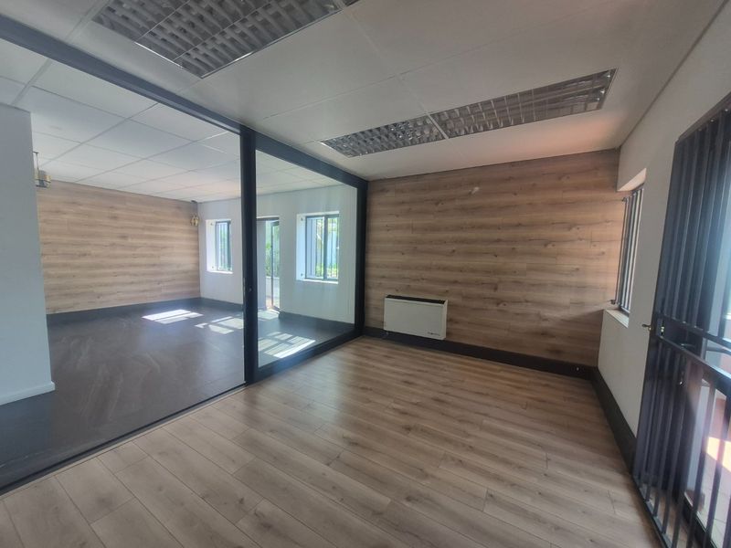 A bright, modern office space featuring wood-look laminate flooring, a glass partition wall, recessed ceiling lighting, and security bars on the windows.