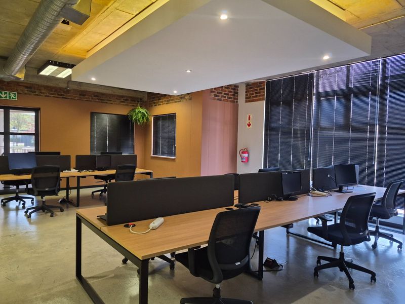Modern open-plan office space featuring exposed concrete ceilings, industrial ducting, exposed brick accents, and polished concrete floors with large windows and venetian blinds.