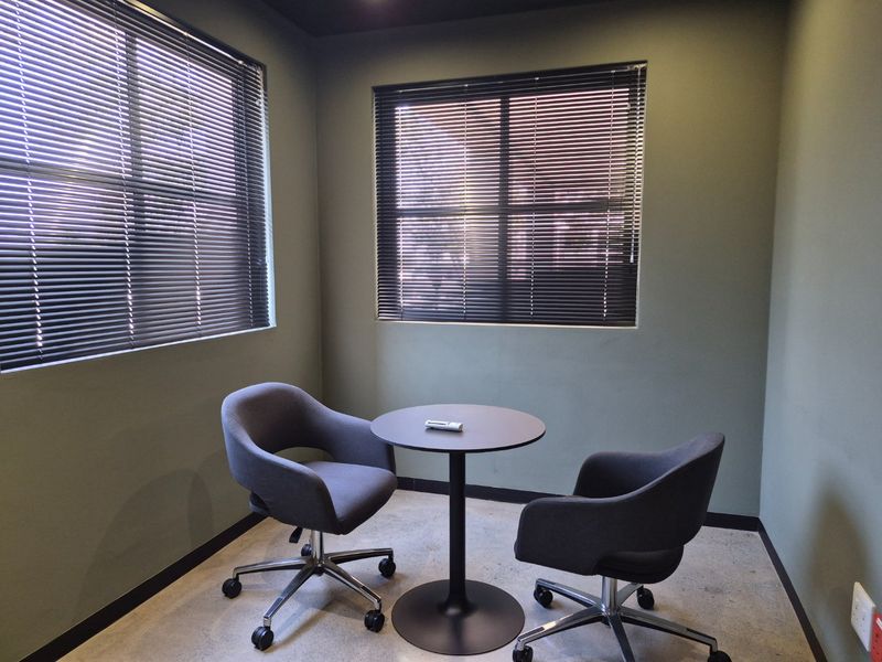 A compact, modern office space featuring polished concrete floors, dark green walls, and dual windows fitted with venetian blinds.