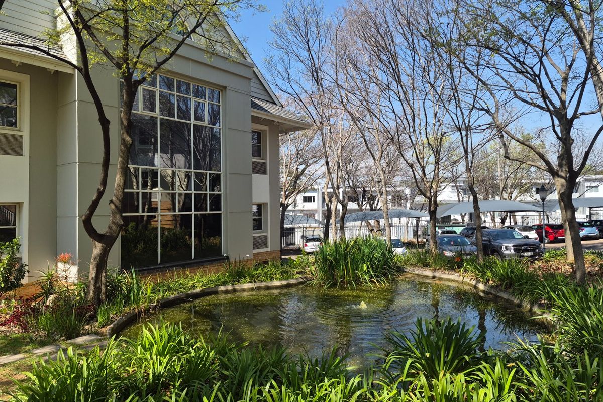 A well-maintained commercial building exterior featuring a landscaped garden with a central pond and fountain, adjacent to a paved parking area.