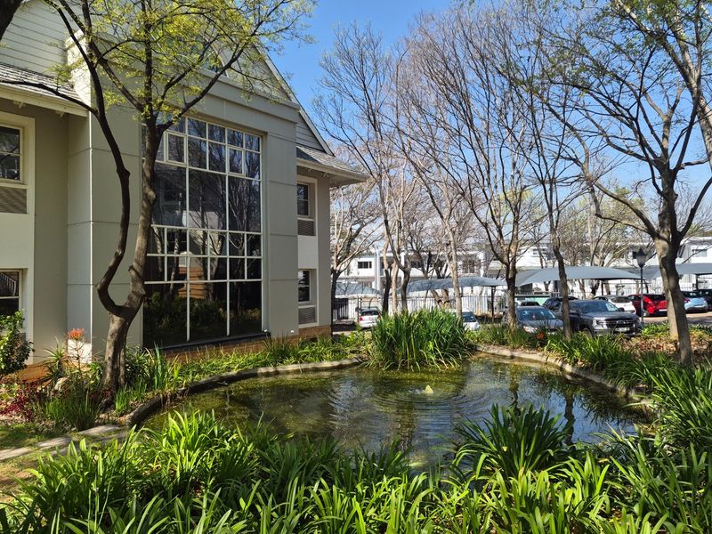 A well-maintained commercial building exterior featuring a landscaped garden with a central pond and fountain, adjacent to a paved parking area.