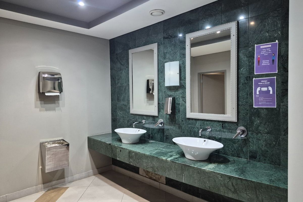A modern commercial bathroom featuring a double vanity with marble countertop, vessel sinks, and wall-mounted faucets against a dark green tiled feature wall.