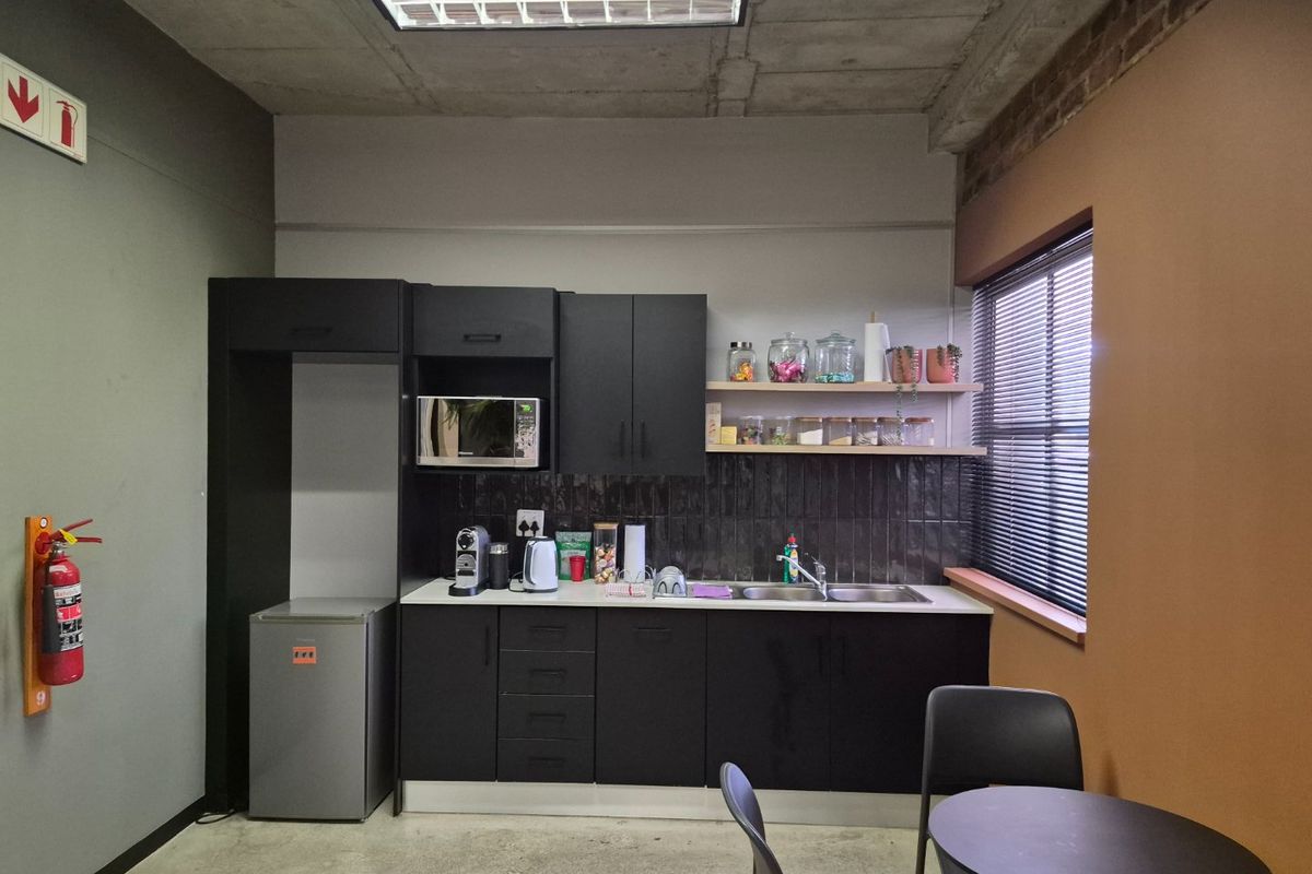 A compact commercial kitchenette featuring modern black cabinetry, a tiled backsplash, stainless steel sink, and industrial-style exposed concrete ceiling.