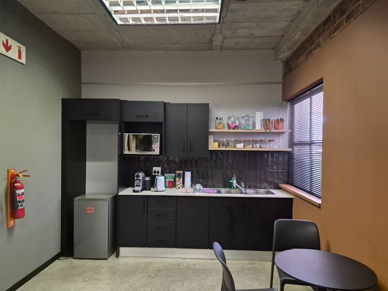 A compact commercial kitchenette featuring modern black cabinetry, a tiled backsplash, stainless steel sink, and industrial-style exposed concrete ceiling.