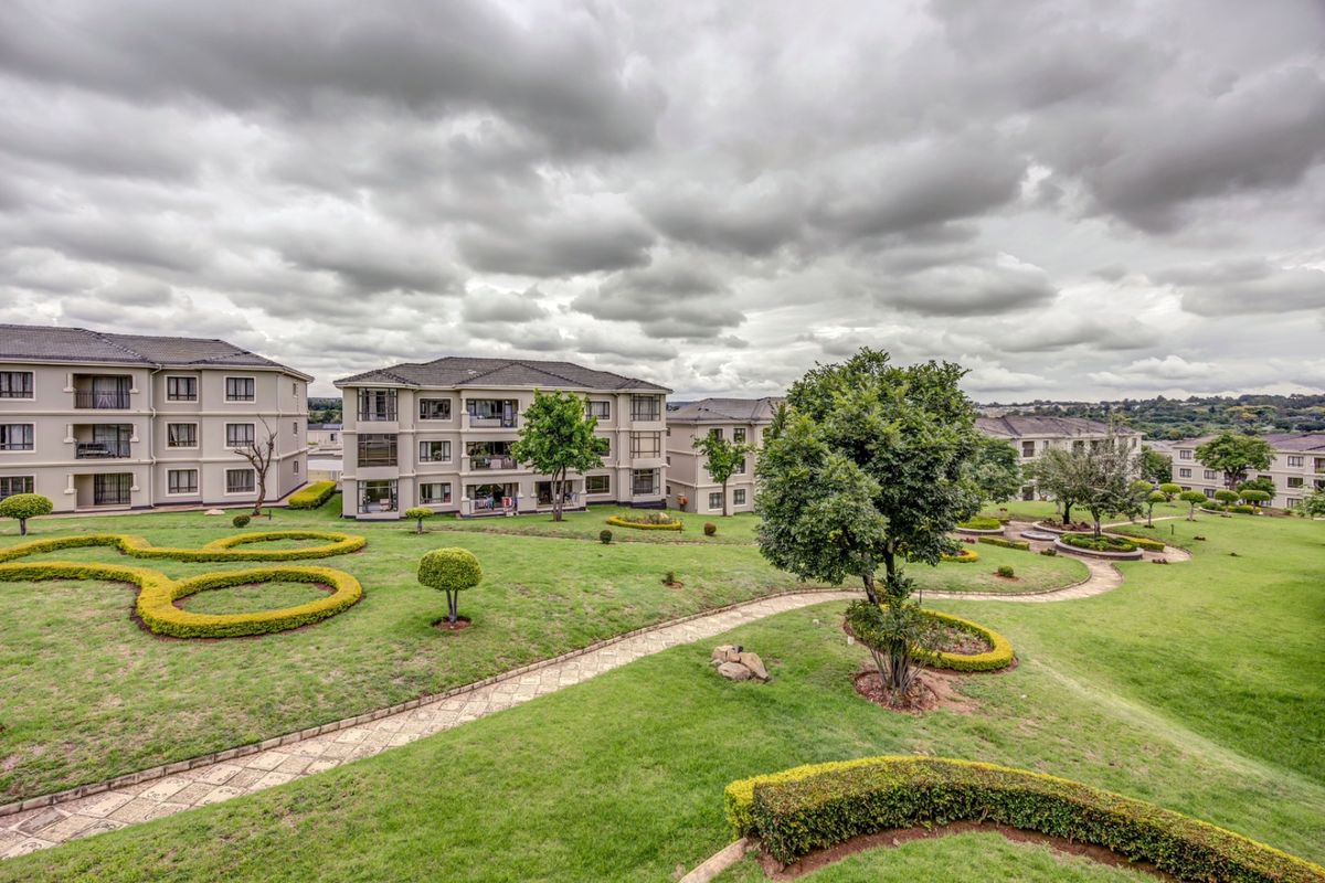 A wide-angle view of the apartment complex grounds, featuring well-maintained landscaped gardens, paved pathways, and multi-story residential buildings under a cloudy sky.