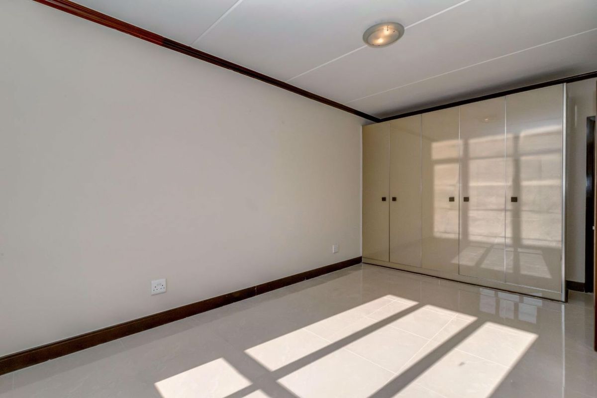 A bright, empty bedroom featuring neutral tiled flooring, built-in cupboards with a glossy finish, and classic ceiling coving.