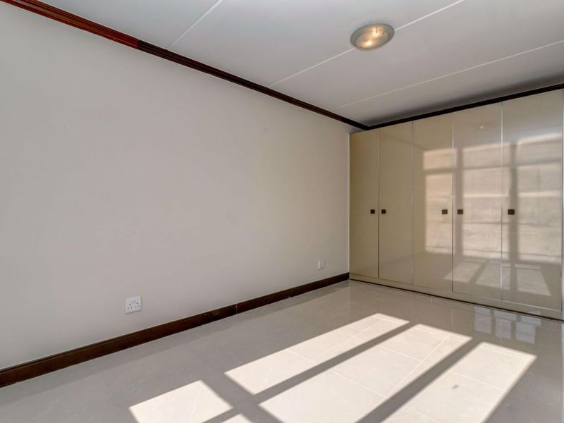 A bright, empty bedroom featuring neutral tiled flooring, built-in cupboards with a glossy finish, and classic ceiling coving.