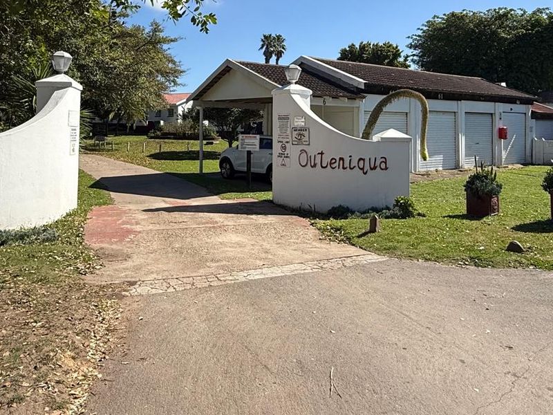 Entrance view of the Outeniqua townhouse complex featuring a gated access point, paved driveway, and covered carport parking area.