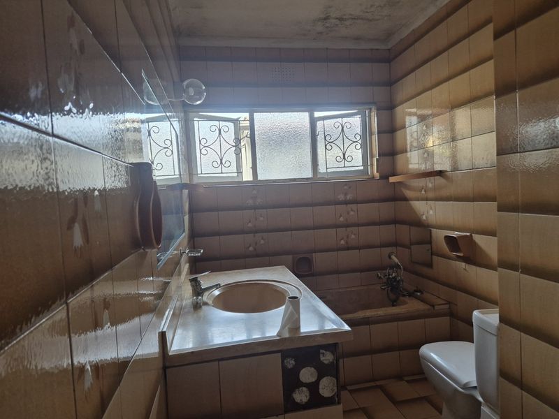 A dated bathroom featuring original tiled walls, a built-in vanity with integrated sink, a bathtub, and a toilet, requiring modernization and cosmetic updates.