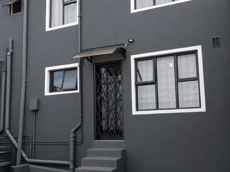 Rear exterior view of the property featuring dark grey plastered walls, aluminium window frames, a security gate, and concrete stairs leading to the entrance.