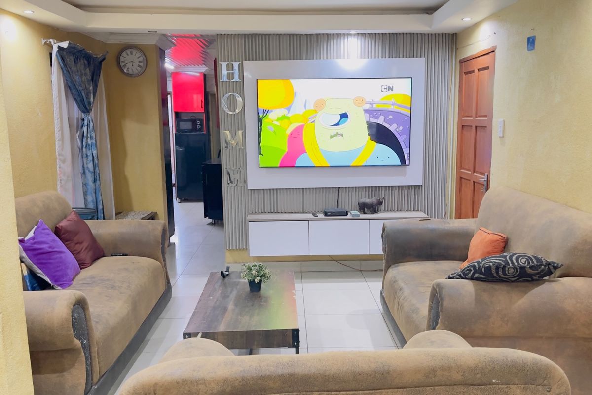 A living room featuring a built-in media wall unit, recessed ceiling lighting, and tiled flooring, with a view into the adjacent kitchen area.