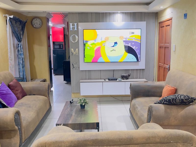 A living room featuring a built-in media wall unit, recessed ceiling lighting, and tiled flooring, with a view into the adjacent kitchen area.
