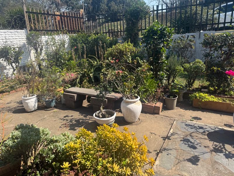 A private garden area featuring concrete paving, established landscaping, and a stone bench, enclosed by perimeter fencing with security wire.