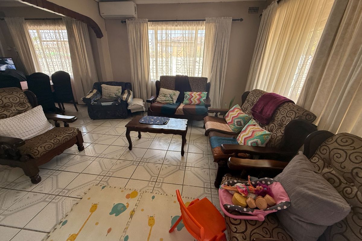A spacious living room featuring ceramic tiled flooring, a wall-mounted air conditioning unit, and large windows fitted with security burglar bars.