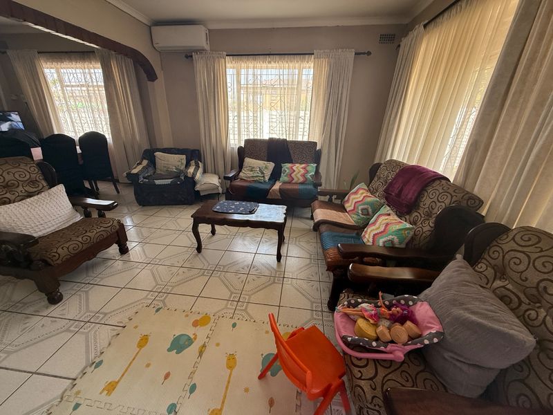 A spacious living room featuring ceramic tiled flooring, a wall-mounted air conditioning unit, and large windows fitted with security burglar bars.