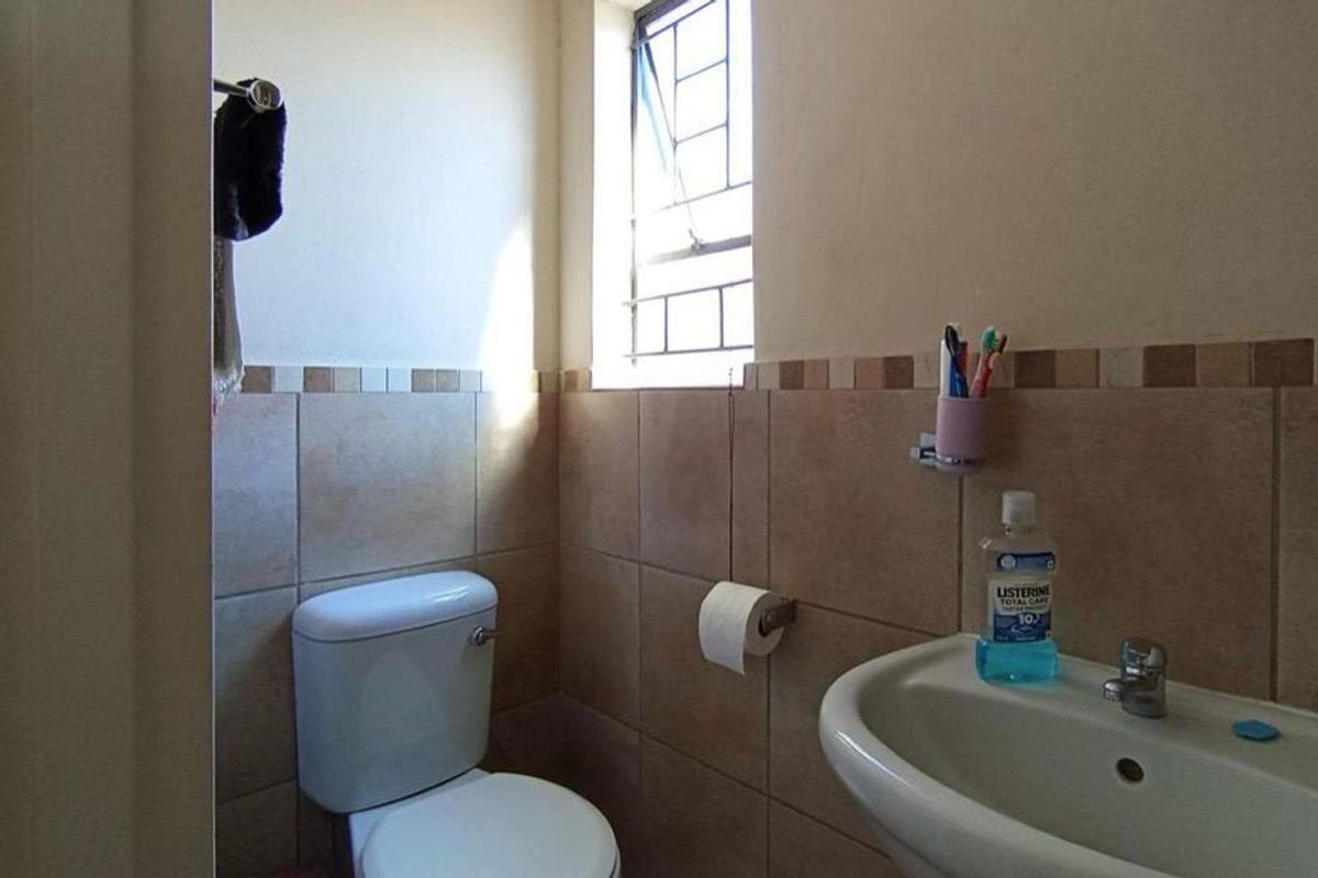 A functional bathroom featuring ceramic wall tiling, a pedestal basin, and a toilet, with natural light provided by a window with security bars.