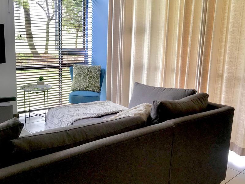 A bright living room featuring large floor-to-ceiling windows with venetian blinds, tiled flooring, and neutral curtain treatments.
