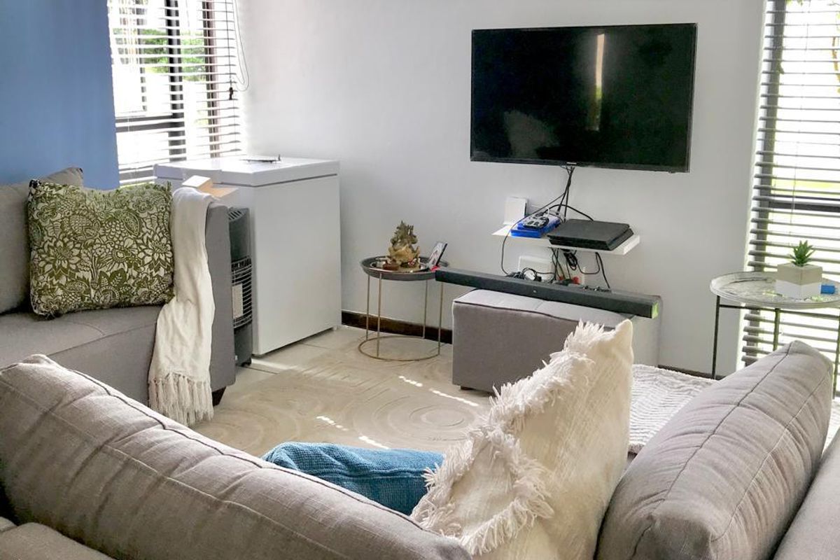 A bright living room featuring large windows with venetian blinds and a wall-mounted television setup.