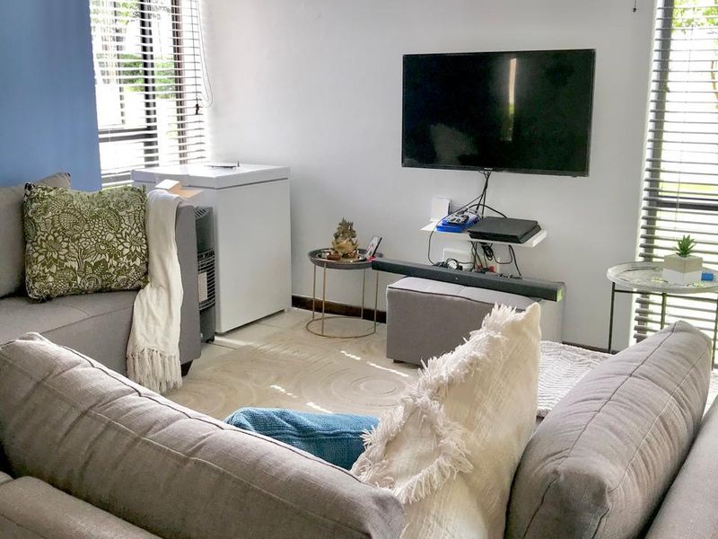 A bright living room featuring large windows with venetian blinds and a wall-mounted television setup.