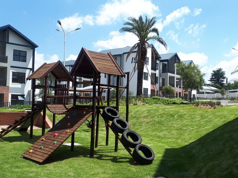 A well-maintained communal garden area featuring a wooden playground structure, manicured lawn, and modern apartment buildings in the background.