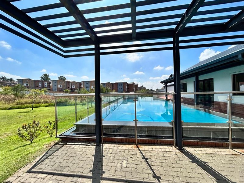 Outdoor pool area featuring a modern pergola, glass balustrade, and paved patio overlooking a landscaped garden and residential complex.