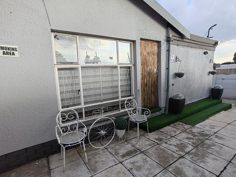 Rear exterior view of the property featuring a paved patio area, artificial grass border, and a wooden entrance door with metal-framed windows.