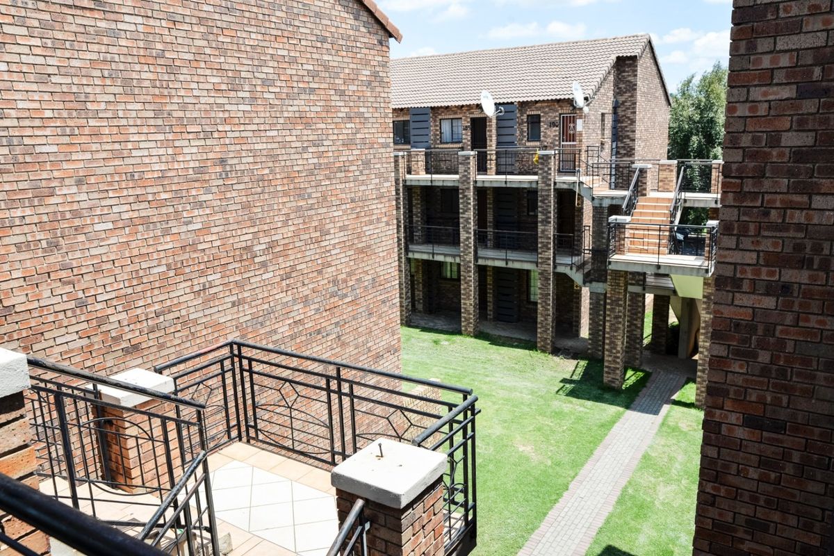 Exterior view of a multi-story brick apartment complex featuring balconies, metal railings, and a shared garden area with paved walkways.