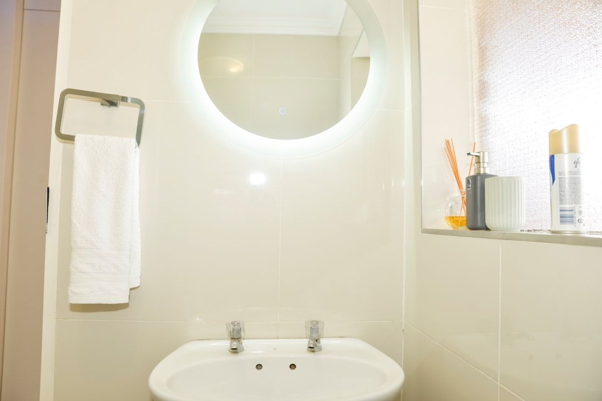 A clean and modern bathroom featuring a pedestal basin, dual tap mixer, and a contemporary backlit circular mirror against neutral tiled walls.