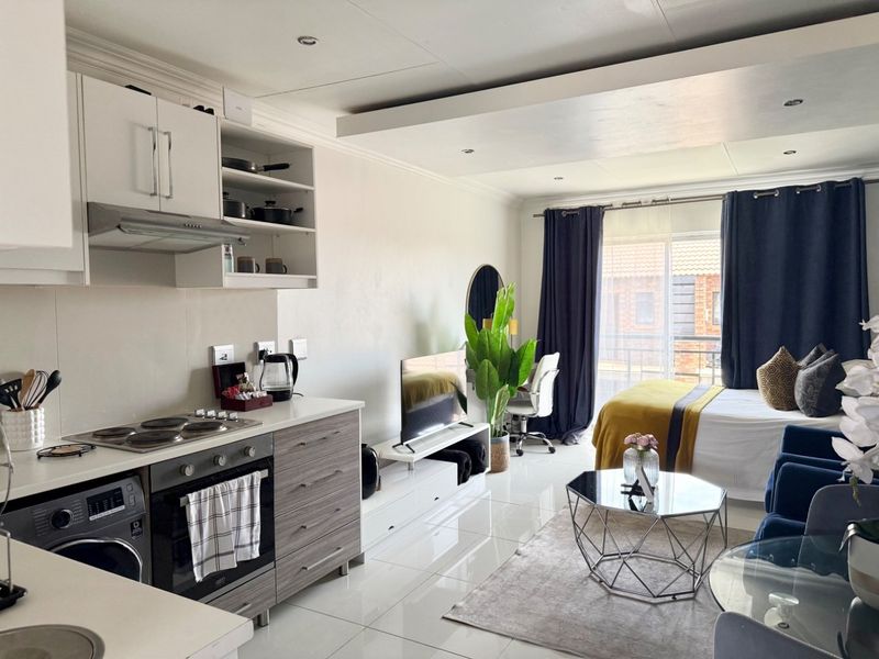 A modern open-plan studio apartment featuring a compact kitchen with built-in appliances and a combined living and sleeping area with access to a balcony.