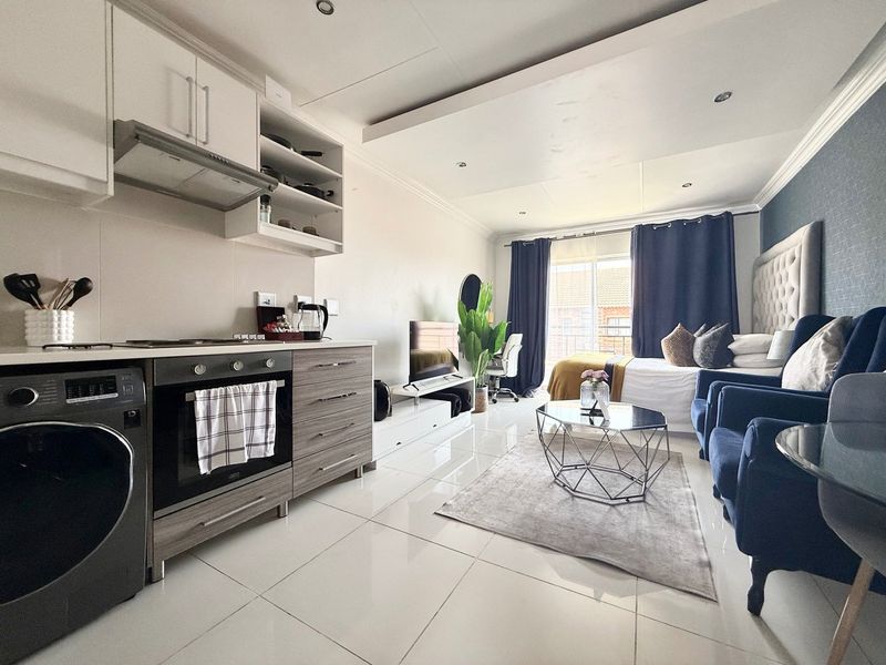 Modern open-plan studio apartment featuring a compact kitchen with built-in appliances, tiled flooring, and a bright living and sleeping area with balcony access.