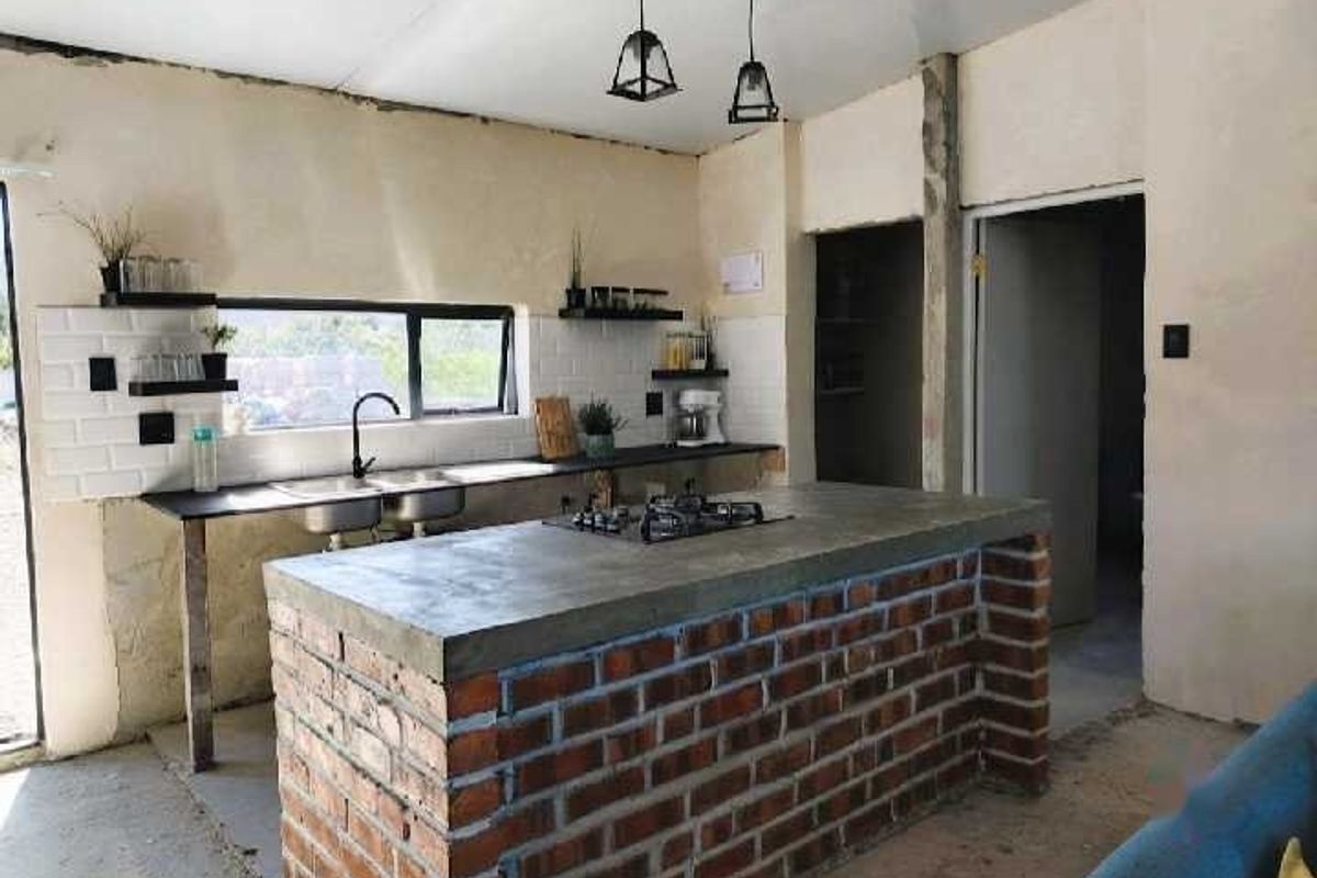 Rustic industrial-style kitchen featuring an exposed brick island with concrete countertop, subway tile backsplash, and double basin sink in an unfinished space.