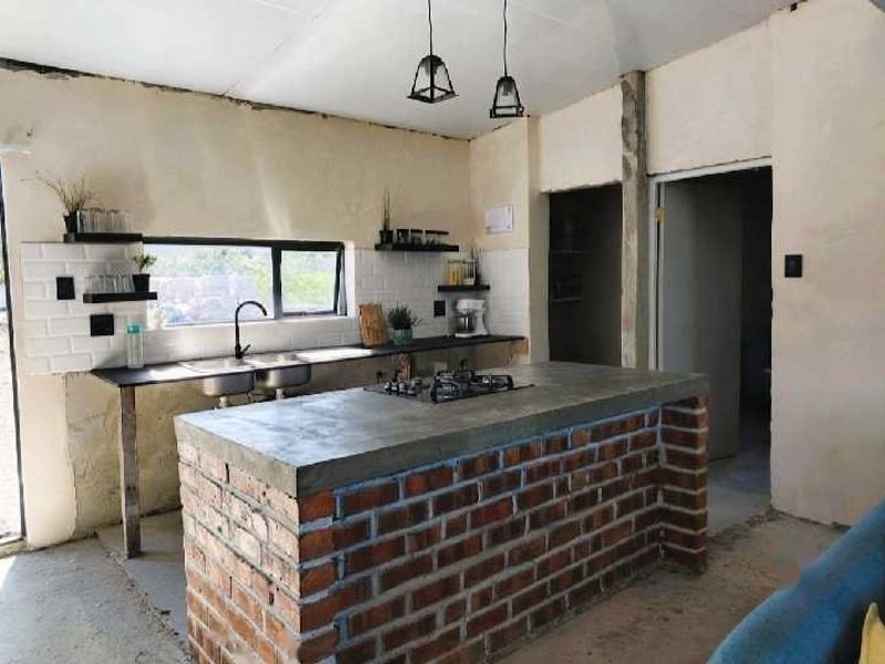 Rustic industrial-style kitchen featuring an exposed brick island with concrete countertop, subway tile backsplash, and double basin sink in an unfinished space.