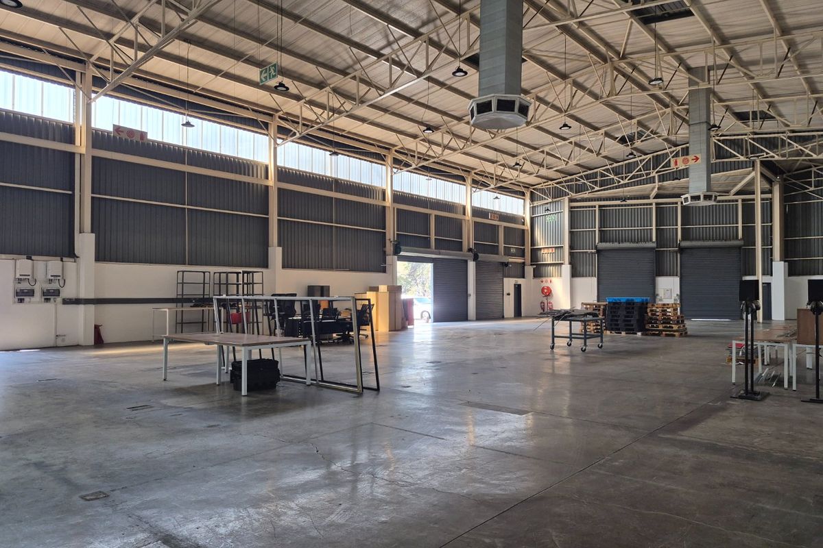 Spacious industrial warehouse featuring high ceilings with exposed steel trusses, concrete flooring, multiple roller shutter doors, and ample natural light from clerestory windows.