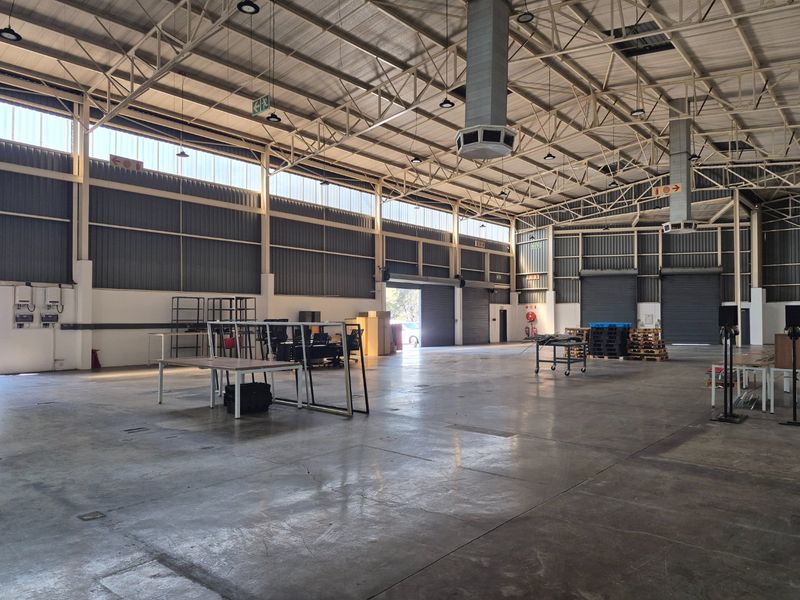 Spacious industrial warehouse featuring high ceilings with exposed steel trusses, concrete flooring, multiple roller shutter doors, and ample natural light from clerestory windows.