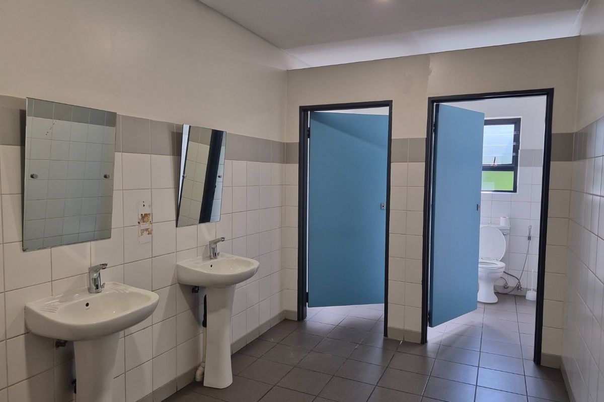 A clean, functional industrial-style communal bathroom featuring dual pedestal washbasins, tiled finishes, and separate toilet cubicles with blue doors.