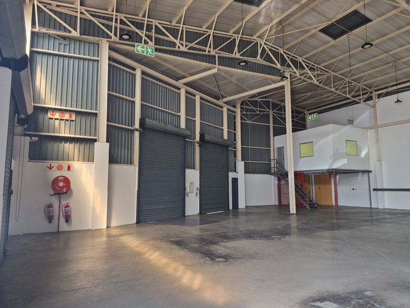 Spacious industrial warehouse featuring high ceilings with exposed steel trusses, dual roller shutter doors, a mezzanine level, and essential fire safety equipment.