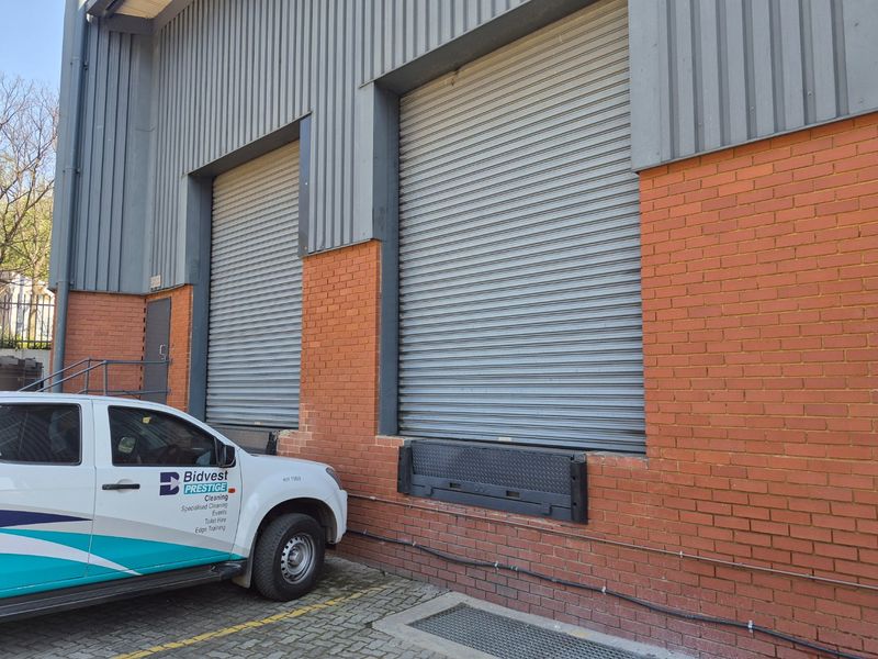 Industrial warehouse exterior featuring two large roller shutter doors, a dock leveler, and a combination of face brick and corrugated metal cladding.