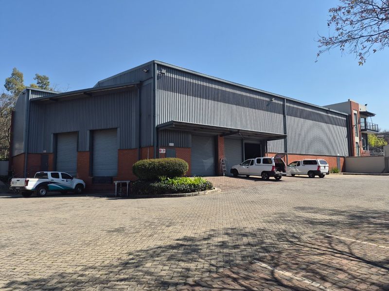 Industrial warehouse facility featuring corrugated metal cladding, brick base, multiple roller shutter doors, and a spacious paved driveway for loading access.