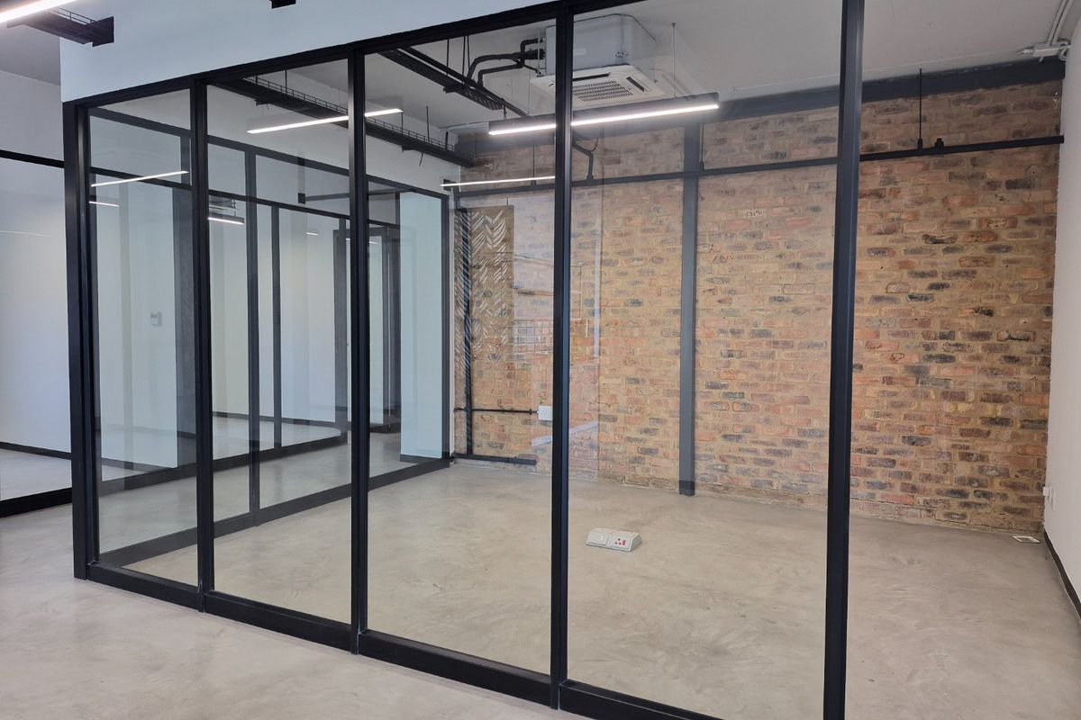 Modern office space featuring industrial-style exposed brick walls, polished concrete flooring, and sleek black-framed glass partitioning with exposed ceiling services.