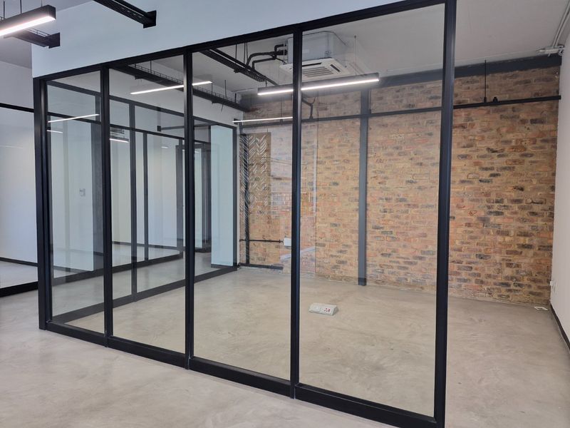 Modern office space featuring industrial-style exposed brick walls, polished concrete flooring, and sleek black-framed glass partitioning with exposed ceiling services.